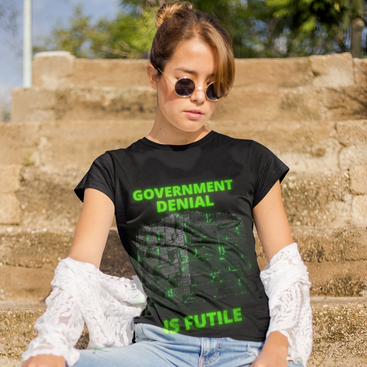 Government Denial Is Futile T-shirt in Black, front print, worn by a model (stone steps, daylight travel). AI-edited photo: the real print artwork was digitally placed on a model photo.