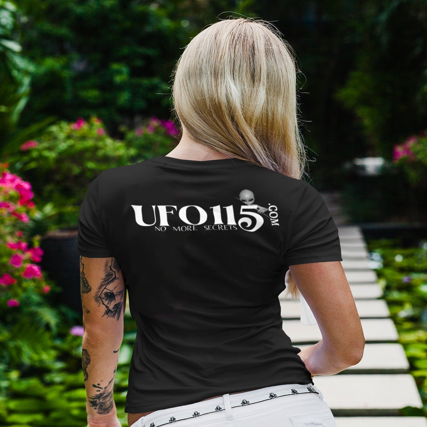 No More Secrets Green Glow Alien Face T-shirt in Black, back print, worn by a model (tropical garden walkway (back)). AI-edited photo: the real print artwork was digitally placed on a model photo.