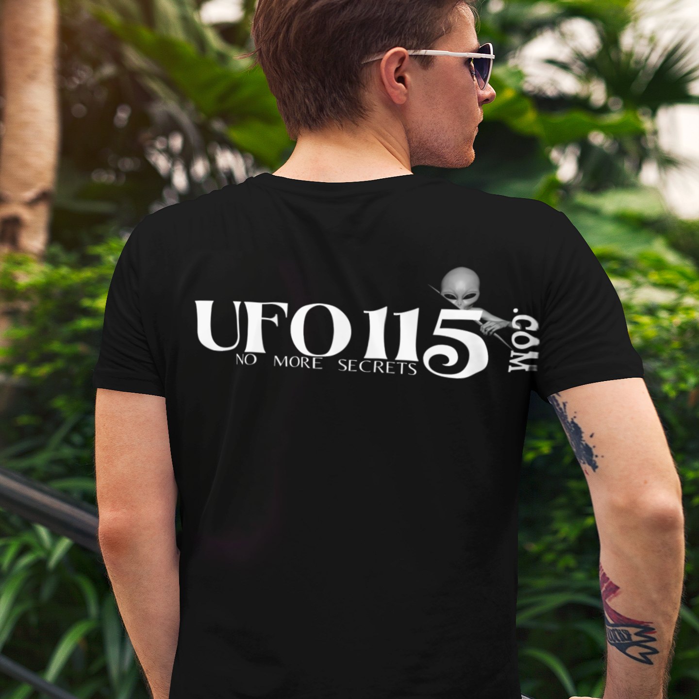 No More Secrets White Alien Face T-shirt in Black, back print, worn by a model (tropical garden, day (back)). AI-edited photo: the real print artwork was digitally placed on a model photo.